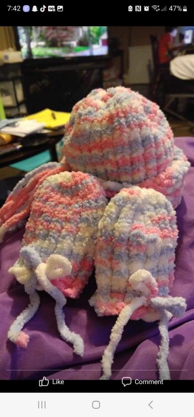 Three handmade knit hats and mittens in pastel colors displayed on a purple surface.