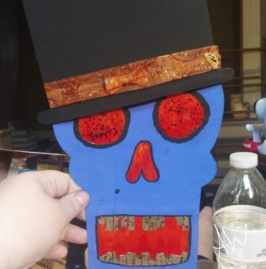 Colorful skull figure with a black top hat and red eyes, held by a hand.